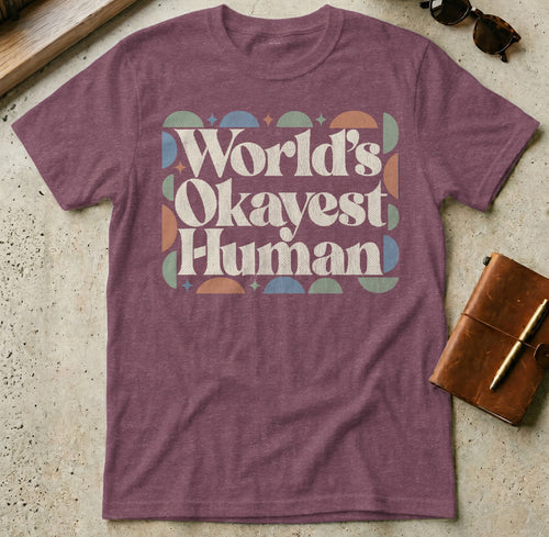 Burgundy t-shirt with "World's Okayest Human" text in colorful geometric shapes.
